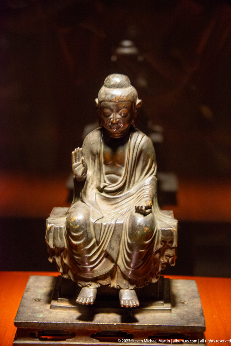 Bronze seated Buddha with one hand raised, the other on the knee, and both feet visible.