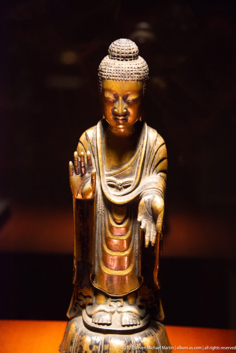 Golden standing Buddha with curled hair, one palm raised, and the other hand extended.