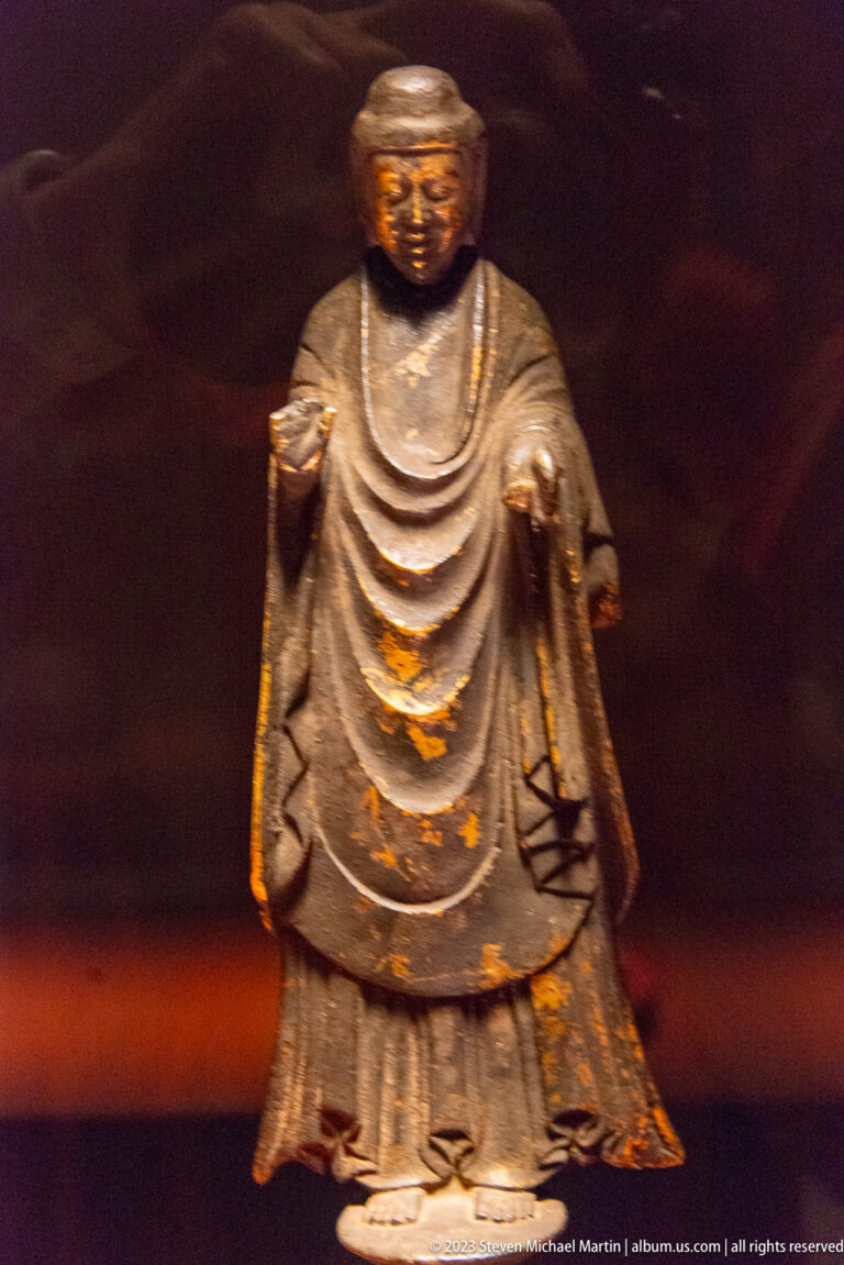 Standing bronze Buddhist figure in broad layered robes with both hands lowered.