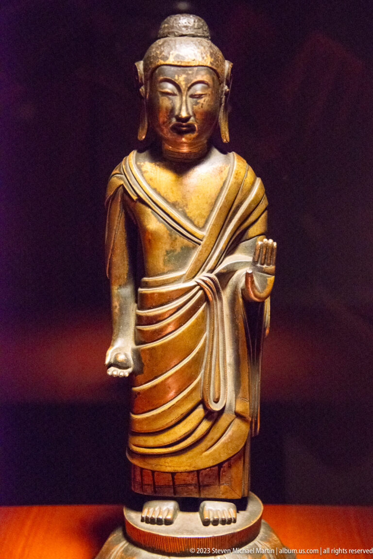 Standing bronze Buddha in a diagonally draped robe with one hand raised near the waist.
