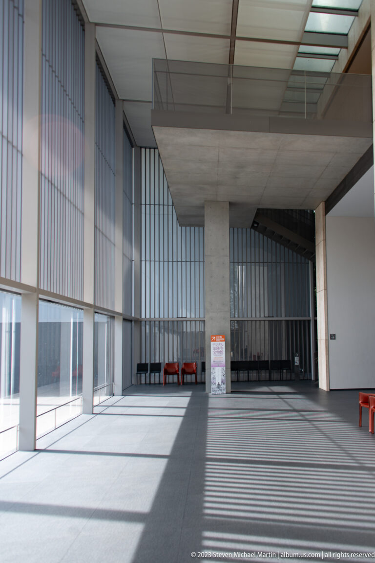 Daylit modern museum hall with vertical louvers, concrete ceiling, glass walls, striped shadows, and red benches.