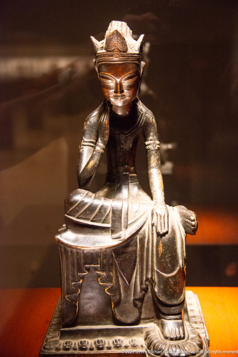 Seated crowned bronze figure with one hand to the cheek and one leg folded across the seat.