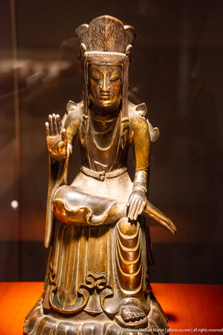 Seated crowned bronze Buddhist figure with one palm raised and a long narrow object behind the body.