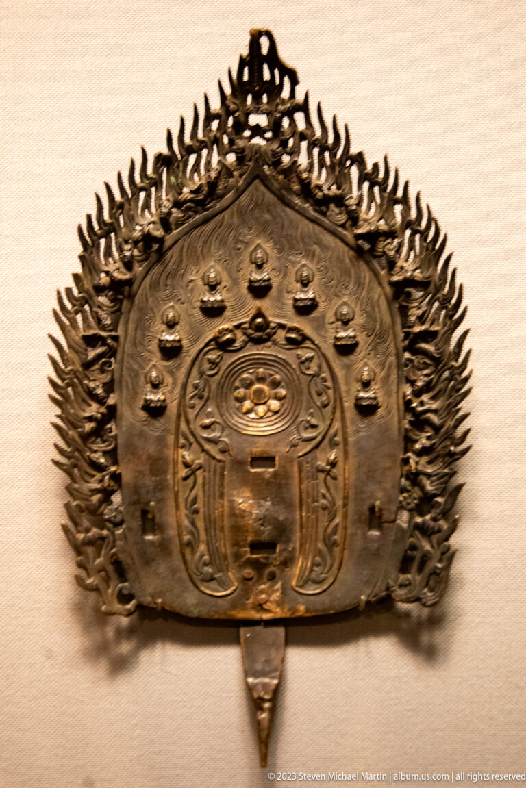 Bronze shrine-shaped Buddhist panel surrounded by radiating flame ornament and small figures.