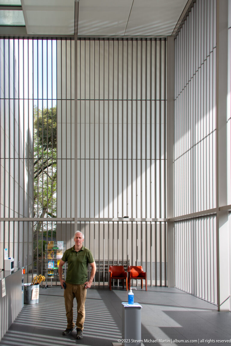 Man seated in a bright museum hall enclosed by tall vertical louvers, with red benches and angled sunlight.