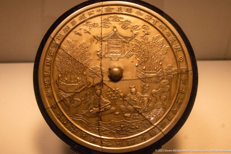 Round bronze mirror back with pavilions, trees, figures, landscape, central knob, and inscription band.