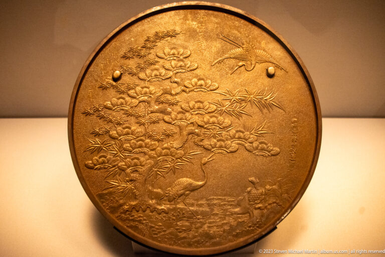 Round bronze mirror back with pine, bamboo, turtle-shaped knob, paired cranes, family crests, and auspicious plants.