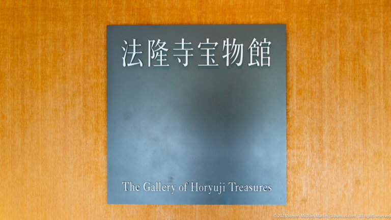 Gray bilingual sign reading The Gallery of Horyuji Treasures mounted on a warm wooden wall.