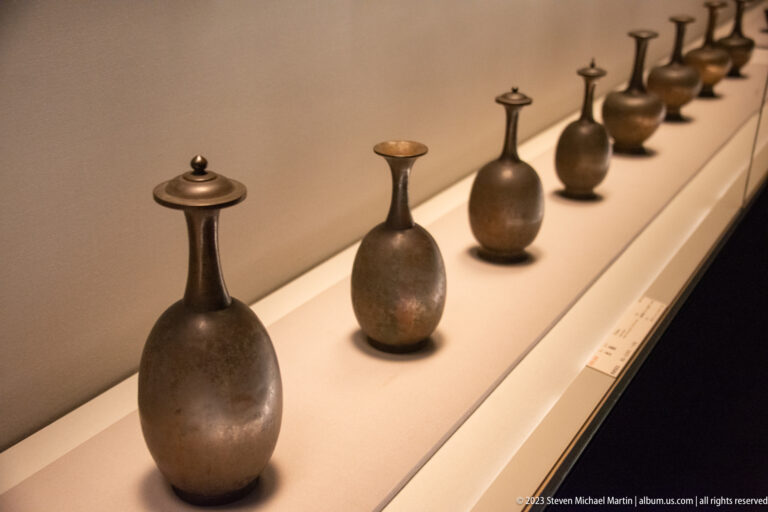 Row of small pear-shaped bronze bottles with narrow necks arranged along a museum shelf.