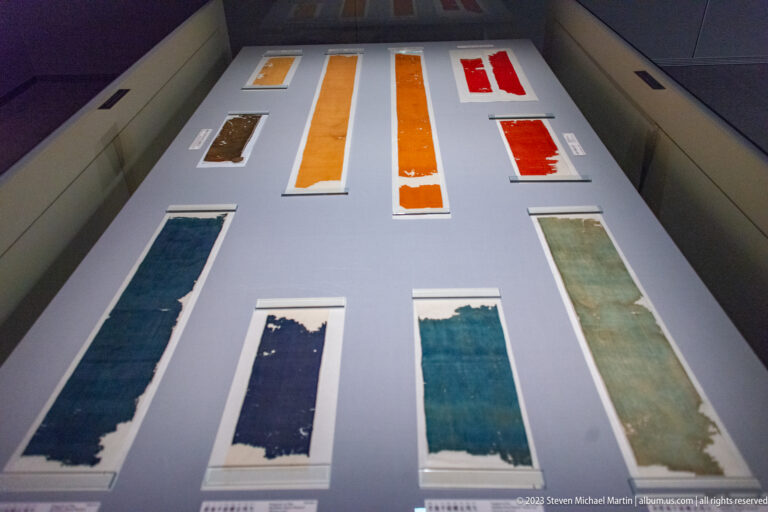 Historic textile fragments in blue, green, orange, and red arranged flat in a museum case.
