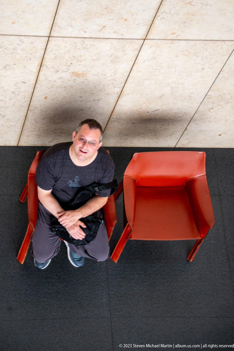 Man seated beside two red chairs, looking upward from a steep overhead viewpoint.