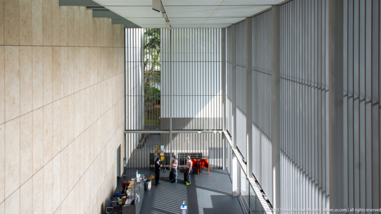 High view into a tall louvered museum hall with visitors, red seating, and striped sunlight below.