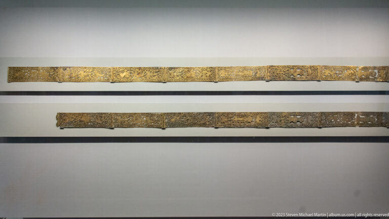 Two long openwork metal fittings with gilded vegetal decoration mounted horizontally in a museum case.