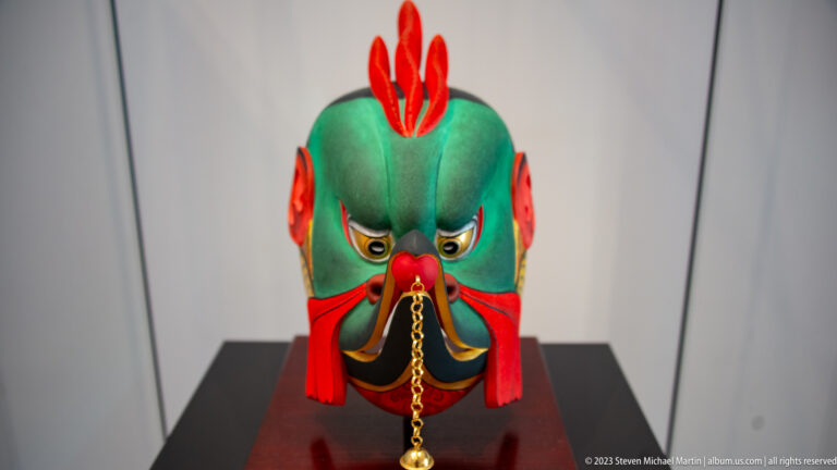 Bright green and red mask with crest, black mustache-like forms, and a dangling gold ornament.