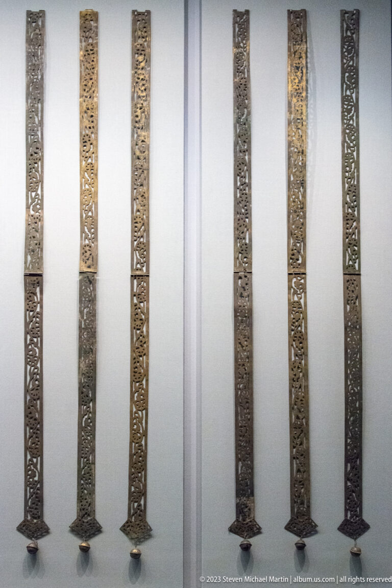 Six tall narrow openwork metal strips displayed vertically on small mounts against a white wall.