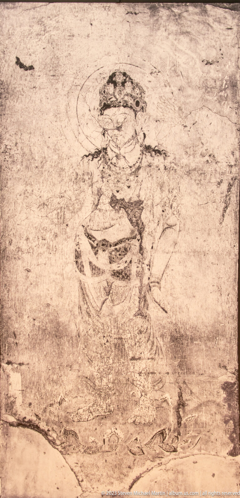 Faint standing haloed Buddhist figure with long robe on a heavily abraded mural.