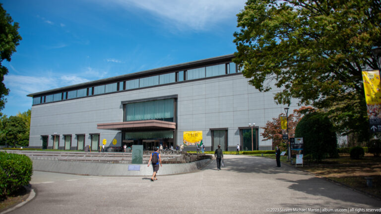 Modern rectangular museum building with pale stone façade, central entrance, clerestory windows, and visitors outside.