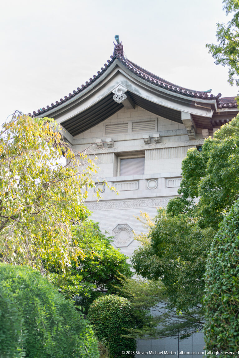 Traditional tiled museum gable and pale stone wall partly hidden by leafy trees.