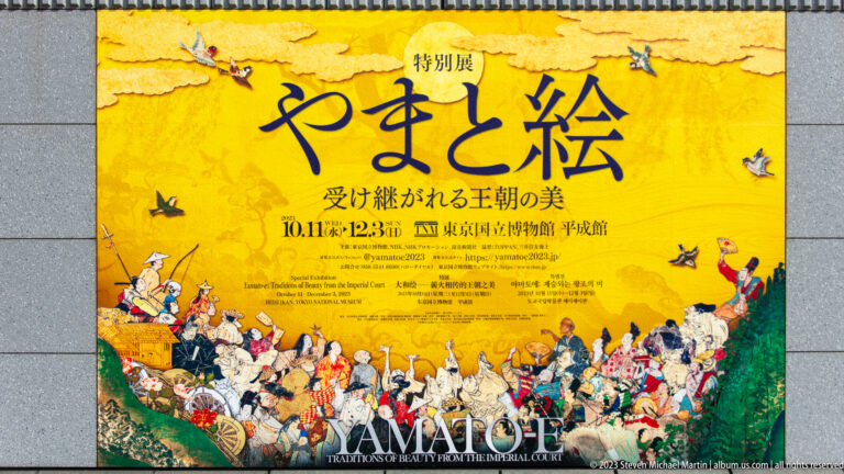 Yellow Yamato-e exhibition poster with large Japanese calligraphy and a procession of historical figures.