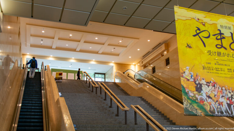Grand museum staircase with visitors, escalators, and a large yellow Yamato-e exhibition banner.