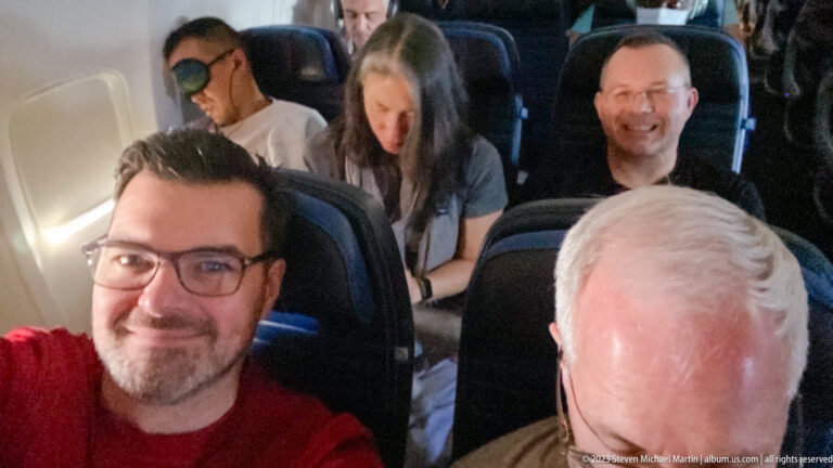 Two male travelers seated in an airplane cabin with other passengers behind them.