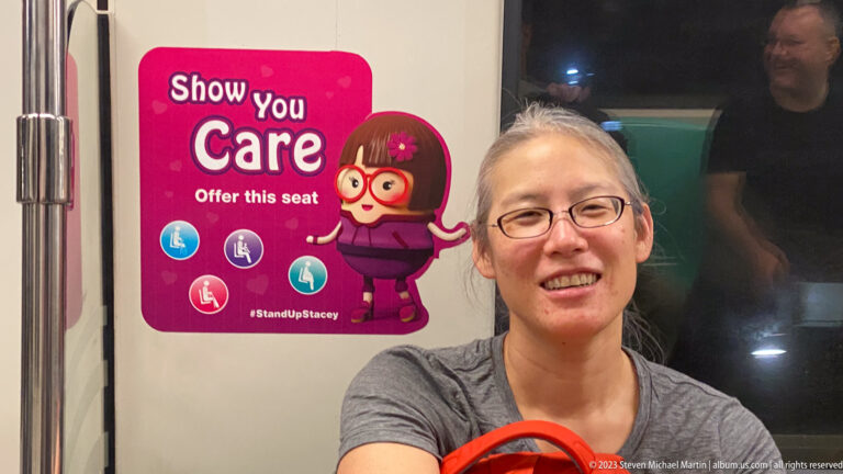 Smiling woman beside a pink transit poster asking riders to offer seats to others.