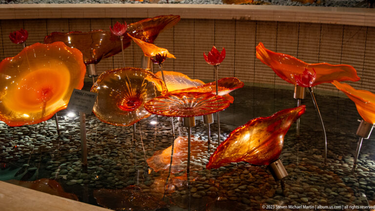 Orange-red glass fish mounted above dark pebbles in an illuminated installation.