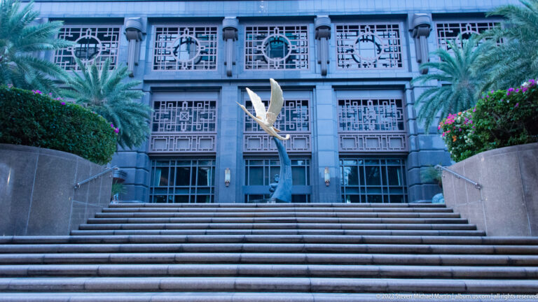 Stylized metal bird sculpture centered before a geometric façade and broad stairs.