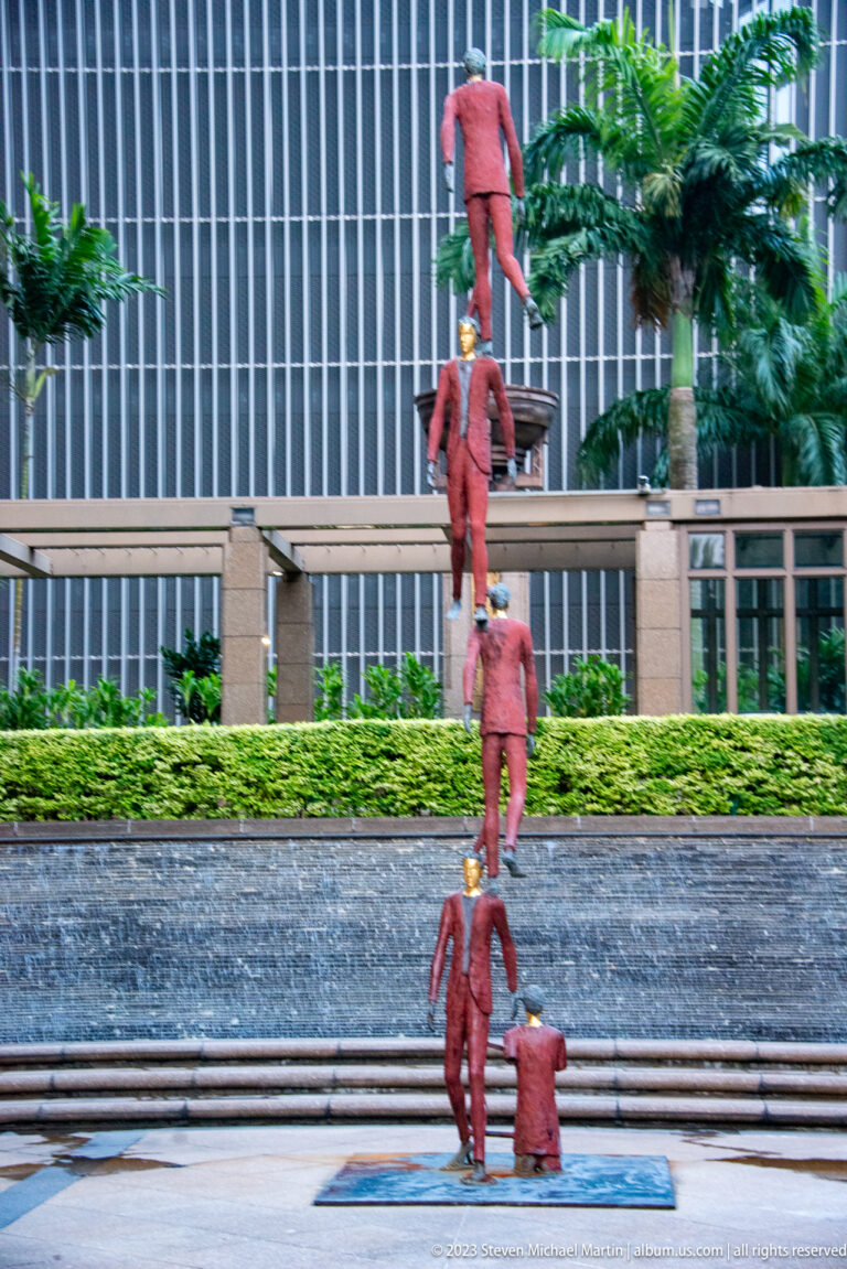 Red human figures stacked vertically in an outdoor fountain before office buildings.