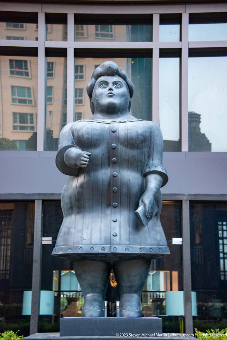 Rounded silver human figure standing outside a modern building with one hand on its hip.