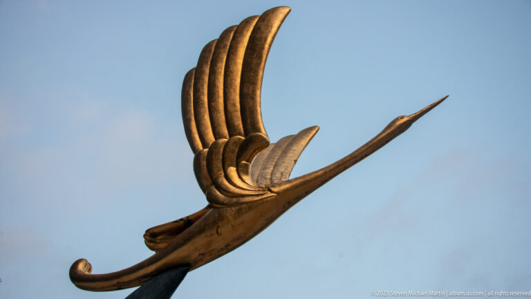 Close-up of a bronze-colored stylized bird sculpture with raised wing against blue sky.