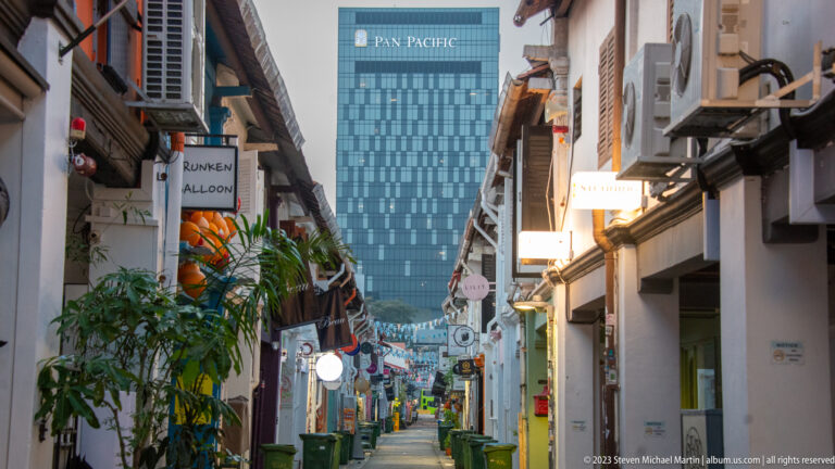 Narrow shophouse lane with signs, plants, air-conditioning units, and a modern tower beyond.