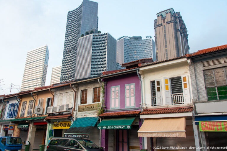 Colorful two-story shophouses with tiled roofs and modern skyscrapers rising behind them.