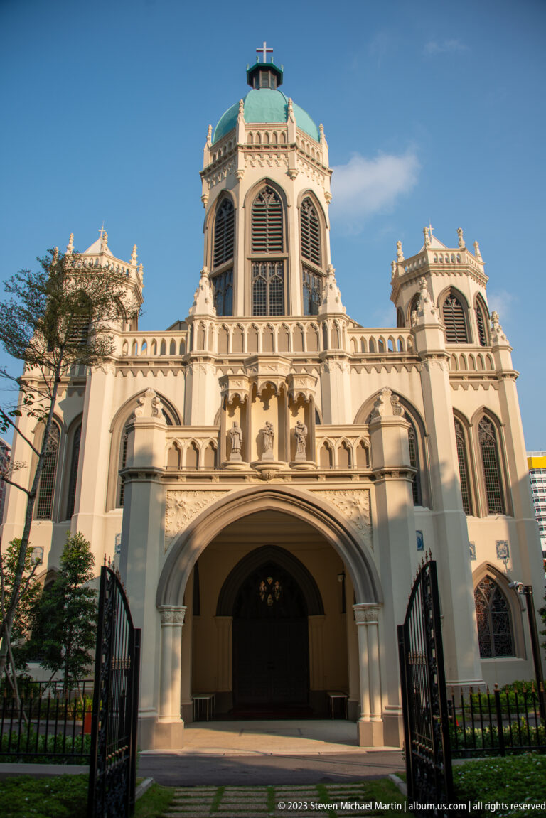White Gothic Revival church with central tower, pointed arches, pinnacles, and open gate.
