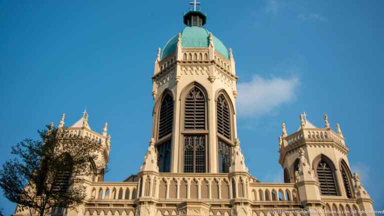 Three pale Gothic Revival church towers with pointed openings and pinnacles against blue sky.