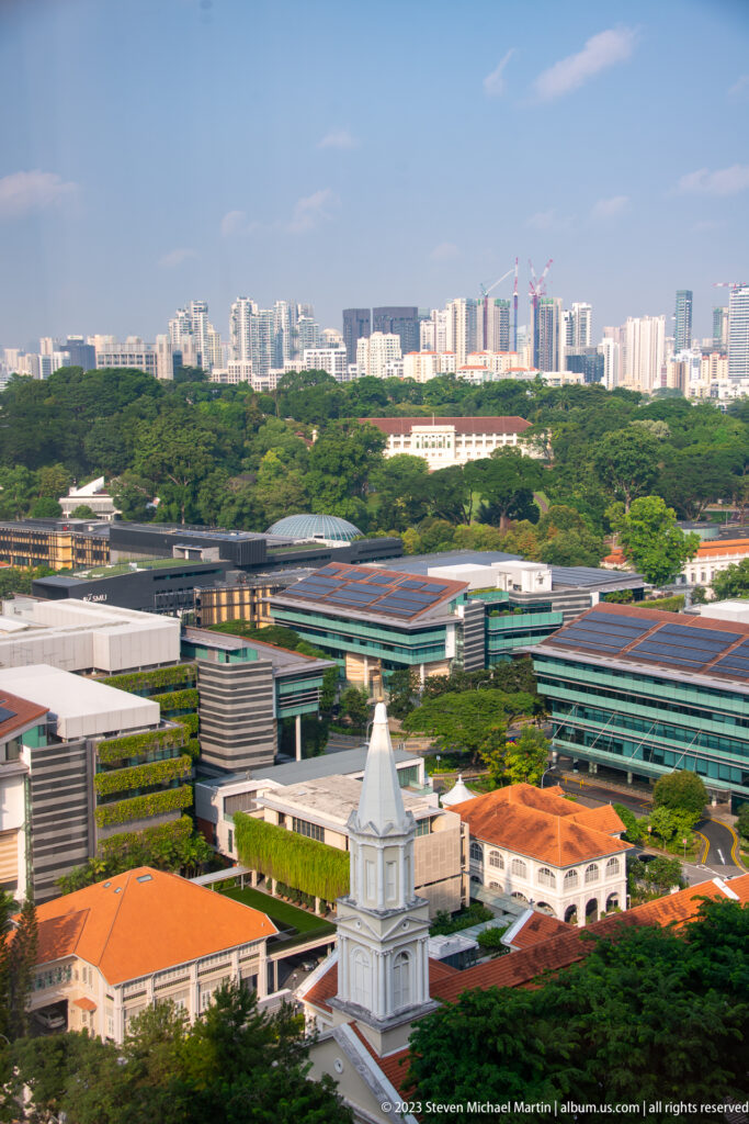 Elevated city view with a white church, red-roofed buildings, greenery, and distant towers.
