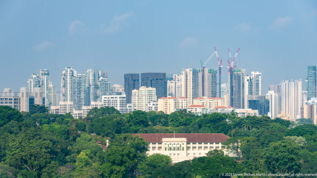 Distant Singapore towers and construction cranes rising beyond dense green trees and low buildings.