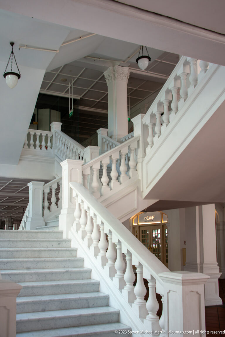 White branching staircase with balustrades, columns, and high ceiling in a colonial-style interior.