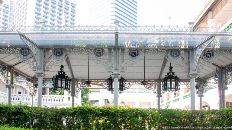 Decorative cast-iron veranda canopy with hanging lanterns, colored glass panels, and hedge below.