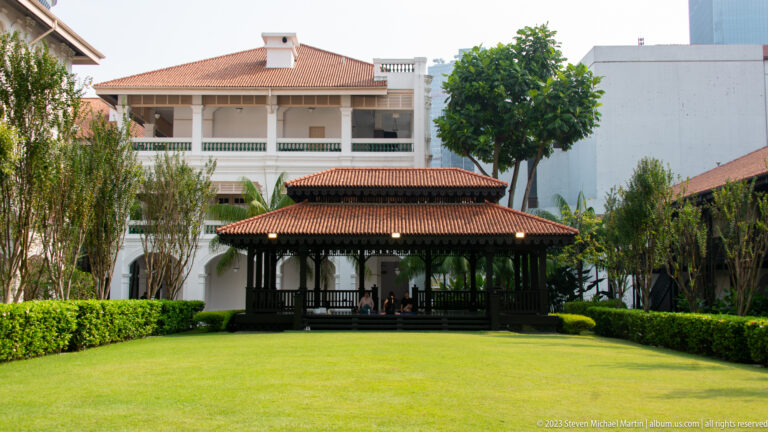 Dark timber garden pavilion on a green lawn amid palms, hedges, and white buildings.