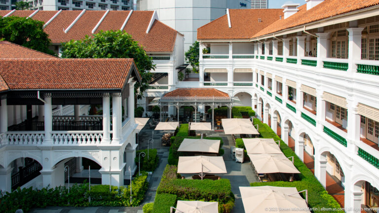 Raffles Hotel courtyard with white arcades, green shutters, red tile roofs, hedges, and café umbrellas.
