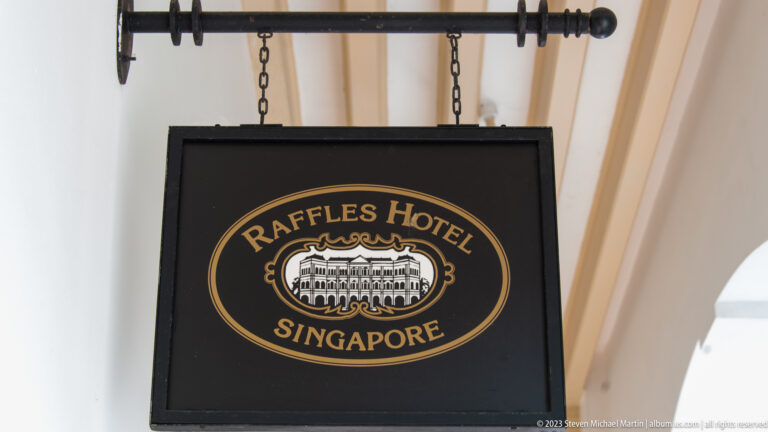 Black hanging sign reading Raffles Hotel Singapore beneath a covered arcade.