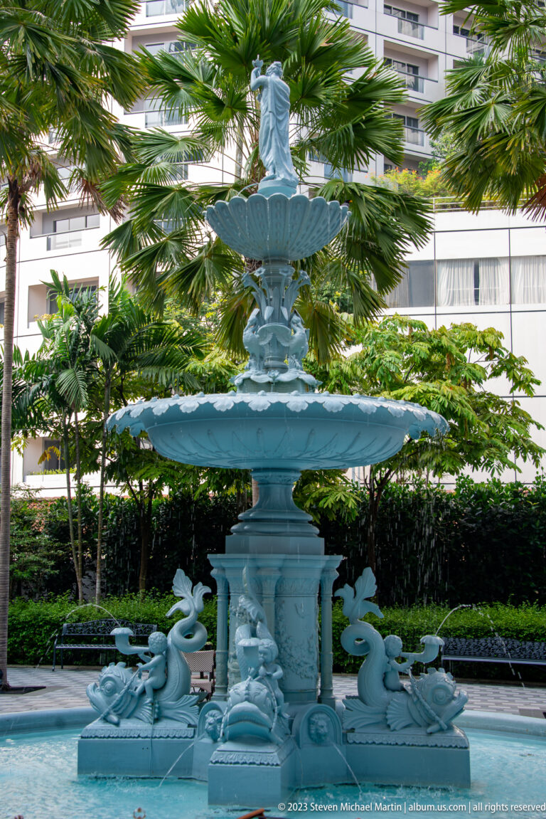 Tiered ornamental fountain with sculpted figures in a palm-lined hotel courtyard.