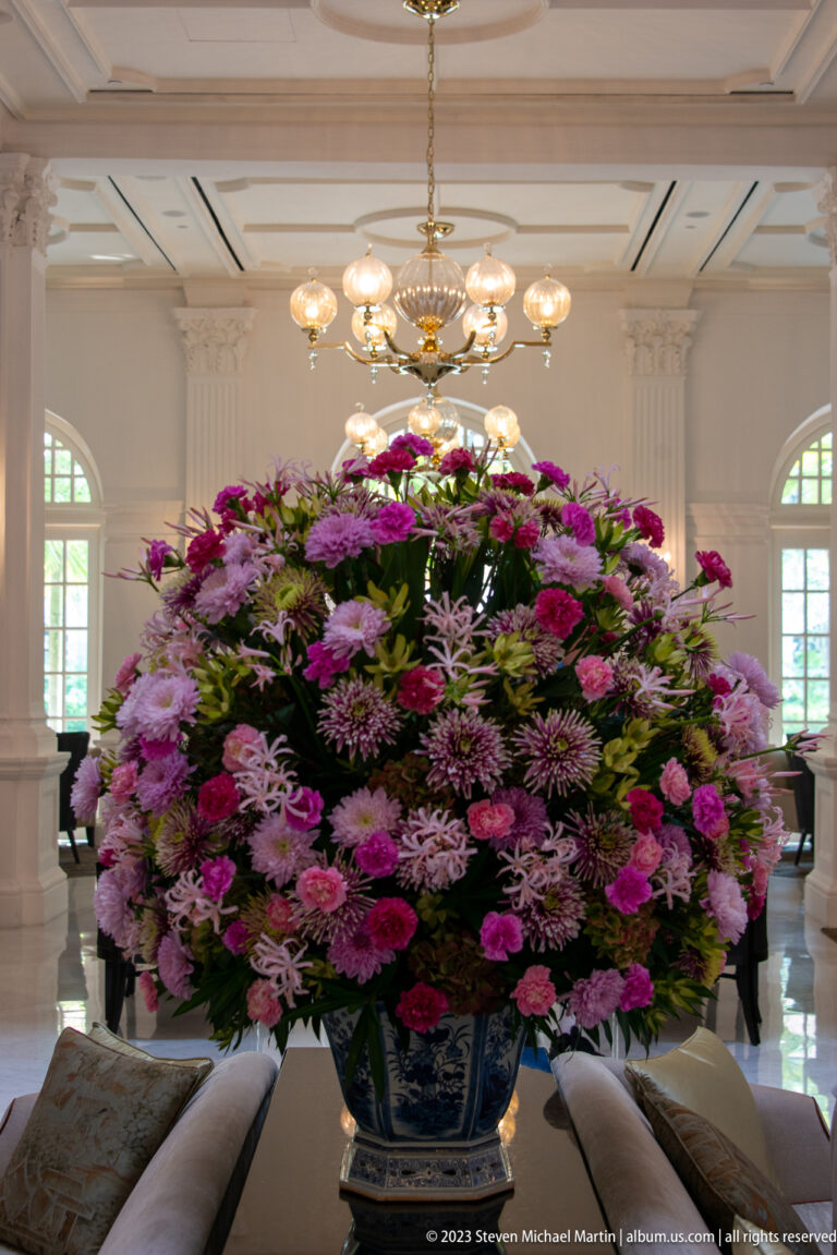 Large pink and purple floral arrangement in a blue-and-white planter beneath chandeliers.