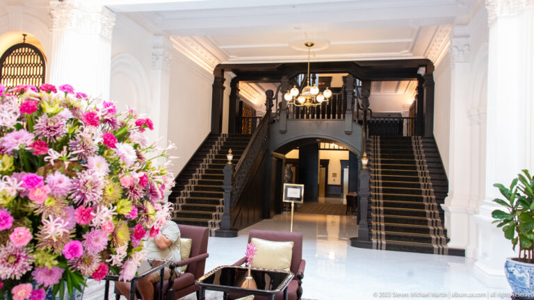 Symmetrical dark-wood staircase rising behind a floral arrangement in a white hotel lobby.