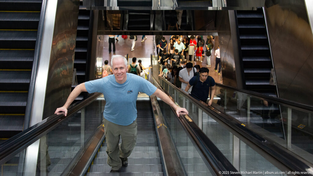 Adult man riding upward on an escalator above a crowded indoor level.