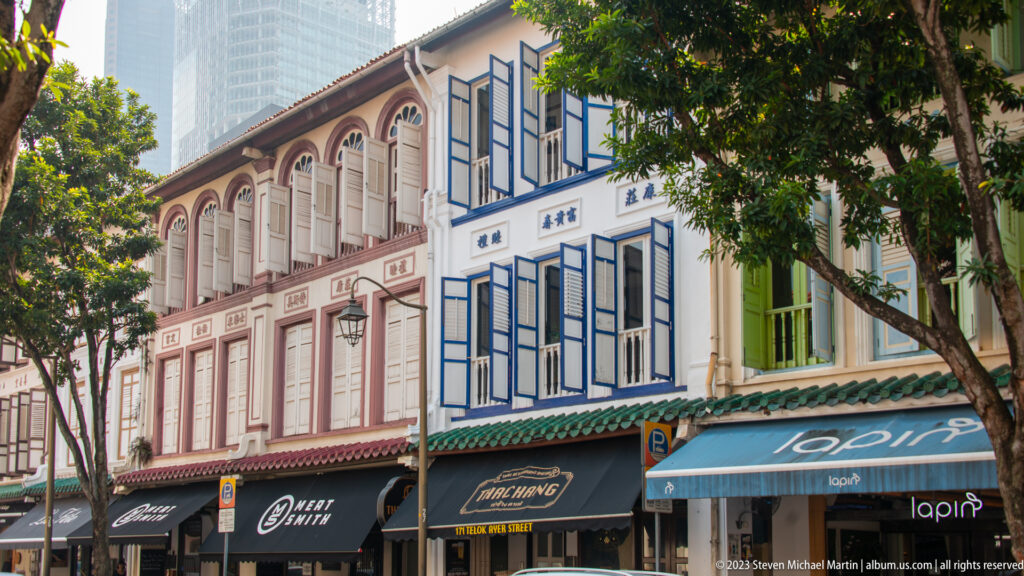 Row of pastel shophouses with arched windows, shutters, awnings, and roadside trees.