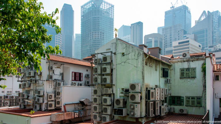 Back walls of low-rise buildings covered with air-conditioning units beneath modern towers.