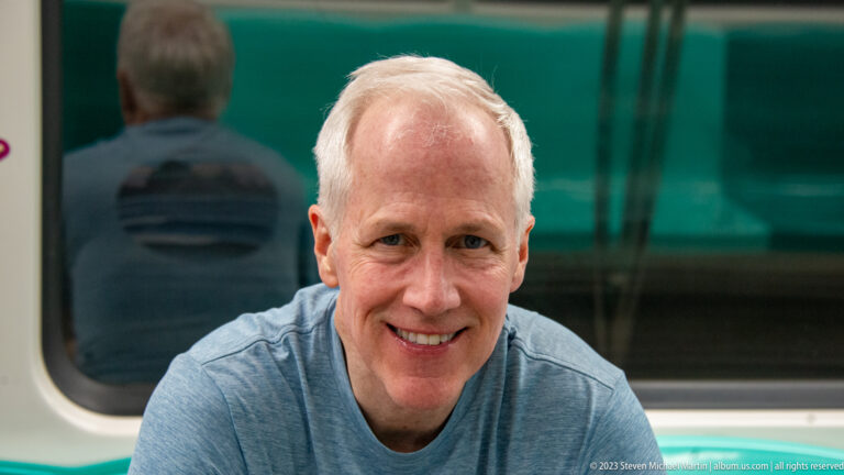 Smiling adult man seated indoors against a teal glass background.