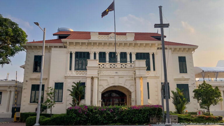 White neoclassical Old Parliament building with green shutters, red roof, and flag.